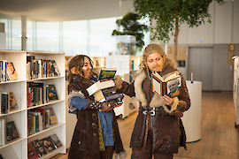 Two people dressed in pseudo-Medieval garb select nine Tolkien-related books in a modern library