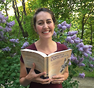 Dr. Sørina Higgins with a copy of her book <span class="push-double"></span>​<span class="pull-double">“</span>The Inklings and King Arthur” in front of trees and a blossoming lilac bush